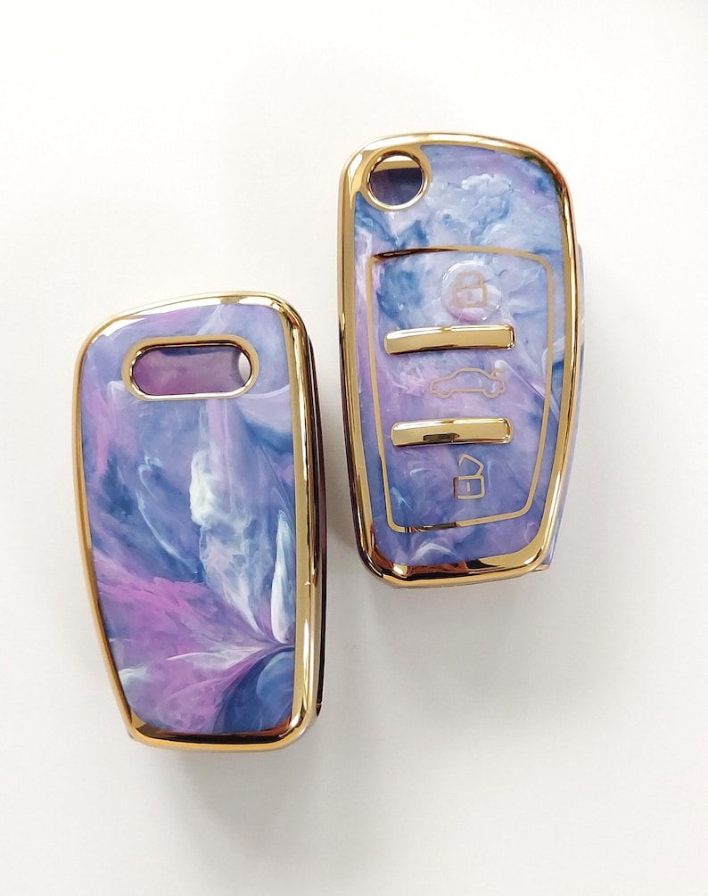 May include: Two car key fobs with a marbled design in shades of purple, blue, and white. The key fobs have a gold-colored trim. One fob has a key ring hole, and the other has buttons for car functions.