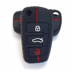 May include: Black car key fob covers with red detailing. The key fobs have a textured honeycomb pattern and feature white and red icons for locking, unlocking, and opening the boot. The key fobs are on a white background.