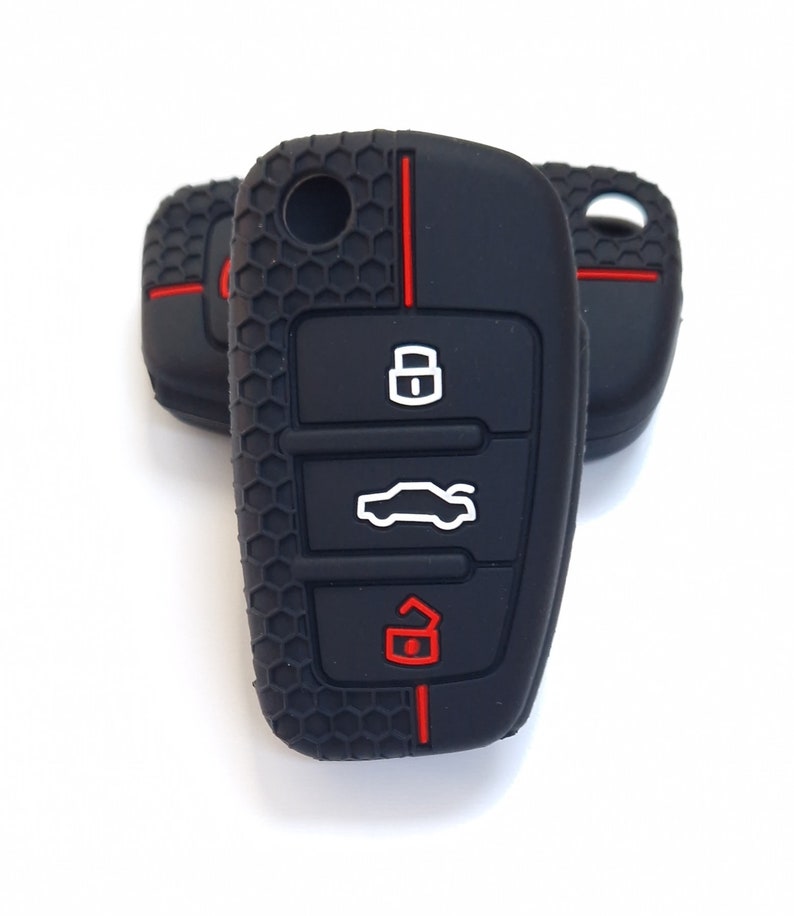 May include: Black car key fob covers with red detailing. The key fobs have a textured honeycomb pattern and feature white and red icons for locking, unlocking, and opening the boot. The key fobs are on a white background.