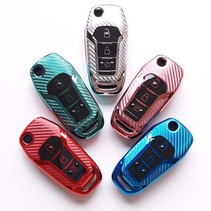 May include: A collection of five car key fobs in various colors, including red, blue, teal, rose gold, and silver. Each fob has a carbon fiber pattern and features buttons for locking and unlocking the vehicle.