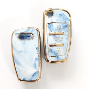 May include: Two car key fobs with a blue and white marbled design. The key fobs have a gold-colored trim. One fob has a key ring hole, and the other has buttons for locking, unlocking, and other car functions.
