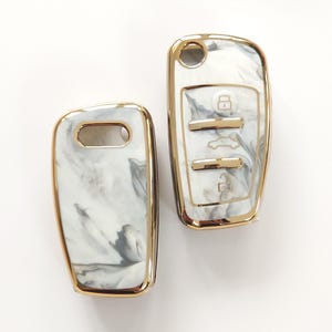 May include: Two car key fobs with a white and gray marble design and gold trim. One fob has buttons with lock, unlock, and trunk release symbols. The other fob has a key ring hole.