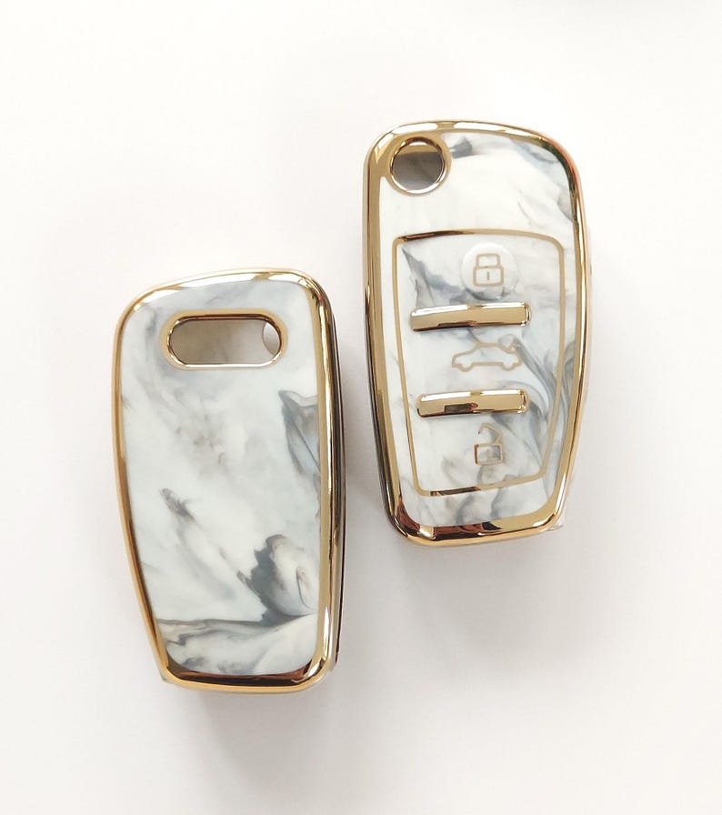 May include: Two car key fobs with a white and gray marble design and gold trim. One fob has buttons with lock, unlock, and trunk release symbols. The other fob has a key ring hole.