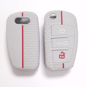 May include: Two-part light grey car key fob cover with red accent stripes. The cover has a honeycomb pattern and embossed icons for lock, unlock, and boot release. The key fob cover is designed to protect the car key.