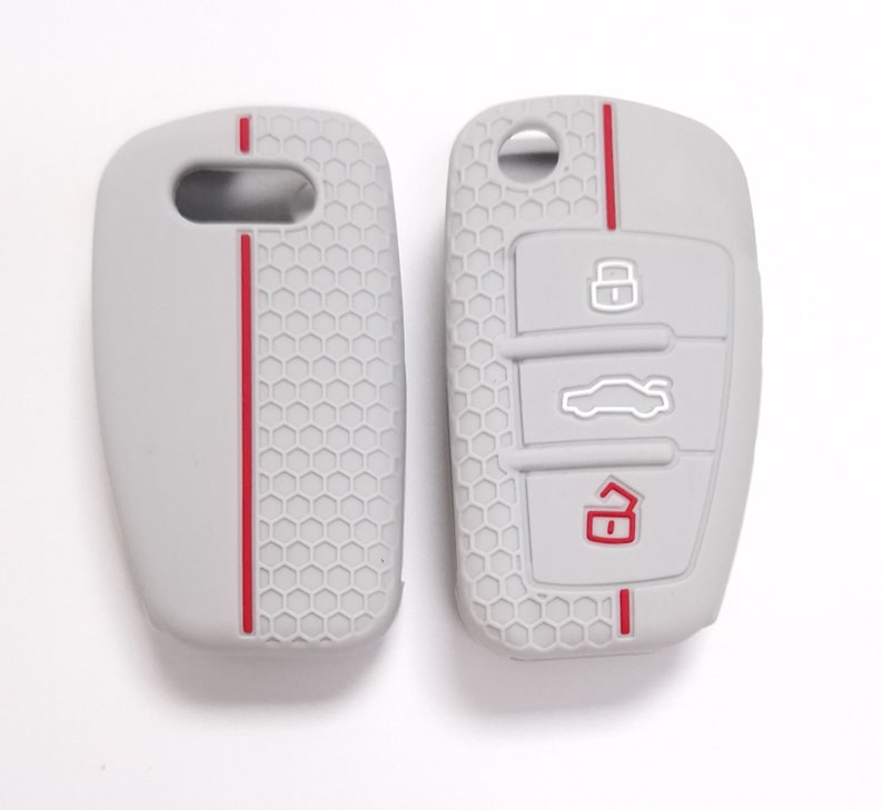 May include: Two-part light grey car key fob cover with red accent stripes. The cover has a honeycomb pattern and embossed icons for lock, unlock, and boot release. The key fob cover is designed to protect the car key.