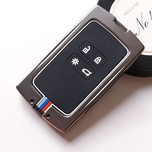 May include: A silver car key fob with four buttons. The buttons are labelled with symbols for unlock, lock, boot, and panic. The fob has a black rubberised surface and a blue, white and red stripe on the side.
