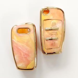 May include: Two car key fobs with a marbled design in shades of peach, yellow, and white. The key fobs have a gold-colored trim and feature key symbols.