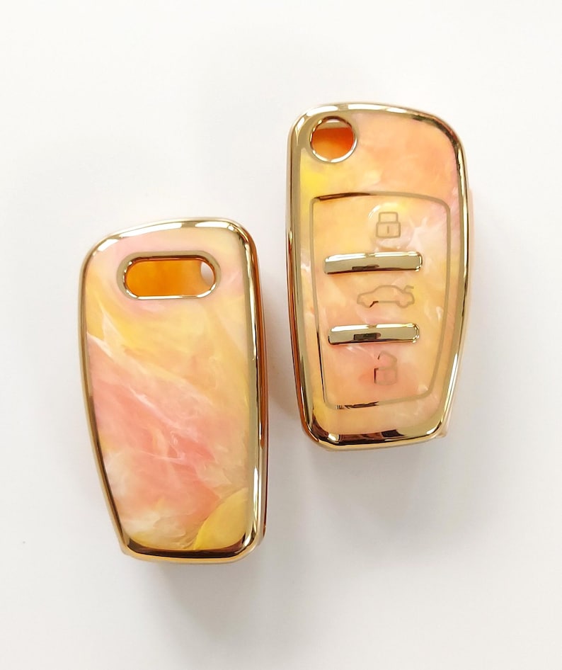 May include: Two car key fobs with a marbled design in shades of peach, yellow, and white. The key fobs have a gold-colored trim and feature key symbols.