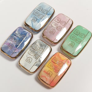 Peut inclure: Six porte-clés de différentes couleurs avec un motif marbré. Chaque porte-clés est doté d'une bordure dorée et présente une série de boutons avec des icônes.