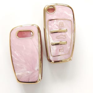 May include: Two car key fobs with a pink marble design and gold-colored trim. The key fobs have a glossy finish and a rectangular shape. One fob has key symbols.