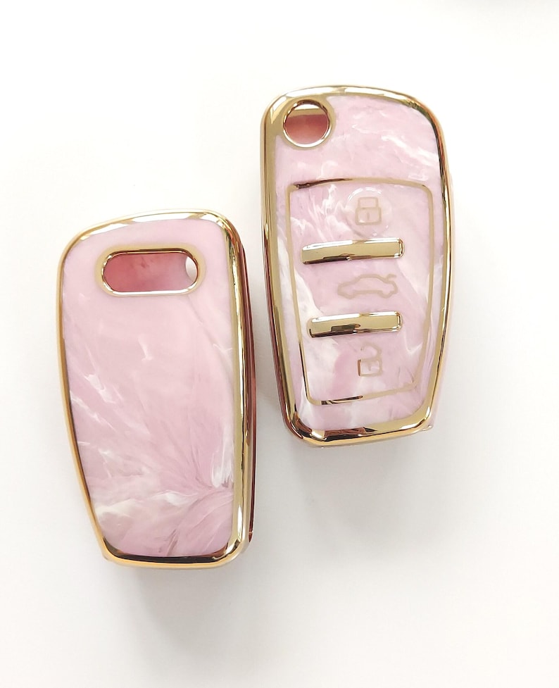 May include: Two car key fobs with a pink marble design and gold-colored trim. The key fobs have a glossy finish and a rectangular shape. One fob has key symbols.