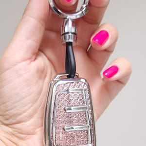 May include: A car key fob with a pink, textured design and silver-colored trim. The key fob is attached to a silver key ring with a black leather strap. The background is a neutral color.