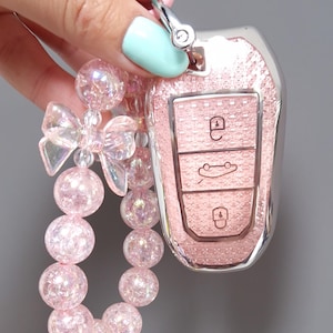 May include: A pink car key fob with a sparkly, textured finish. The fob has three button icons and is attached to a pink beaded keychain with a bow and iridescent beads. The keychain is held by a hand with light blue nail polish.