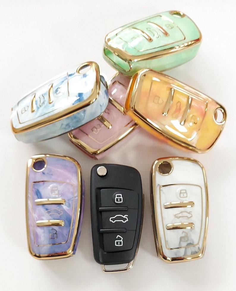 May include: A collection of car key fobs and key covers in various colors, including green, orange, pink, blue, purple, and white. The key covers have a marbled design with gold-colored trim. One black car key is also present.