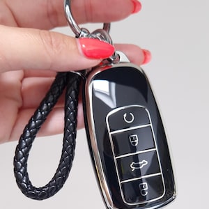 以下が含まれることがあります： シルバーのトリムと黒い編み込みレザーキーチェーンが付いた黒い車のキーフォブ。キーフォブには、ロック、ロック解除、トランクオープンの記号が付いた3つのボタンがあります。キーリングはシルバーです。