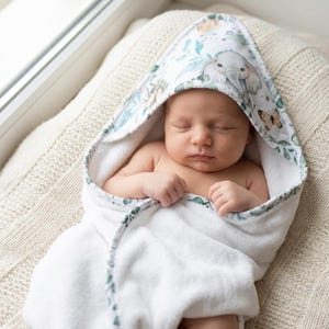 Op de afbeelding: Een slapende baby gewikkeld in een witte badhanddoek met capuchon. De capuchon heeft een aquarelontwerp van een olifant, vlinders en bladeren in groen- en bruintinten. De handdoek is zacht en pluizig, perfect na het bad.
