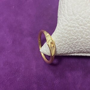 May include: A gold ring featuring a small, round gemstone. The ring has a smooth, polished band and a simple design. It is displayed on a white surface against a purple background.