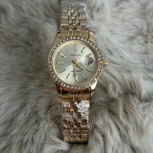 Puede incluir: Reloj de pulsera dorado con esfera redonda y correa met&aacute;lica. La esfera est&aacute; rodeada de peque&ntilde;os cristales transparentes. La esfera del reloj tiene la palabra "COLISEUM" impresa. El reloj a&uacute;n est&aacute; envuelto en pl&aacute;stico protector.