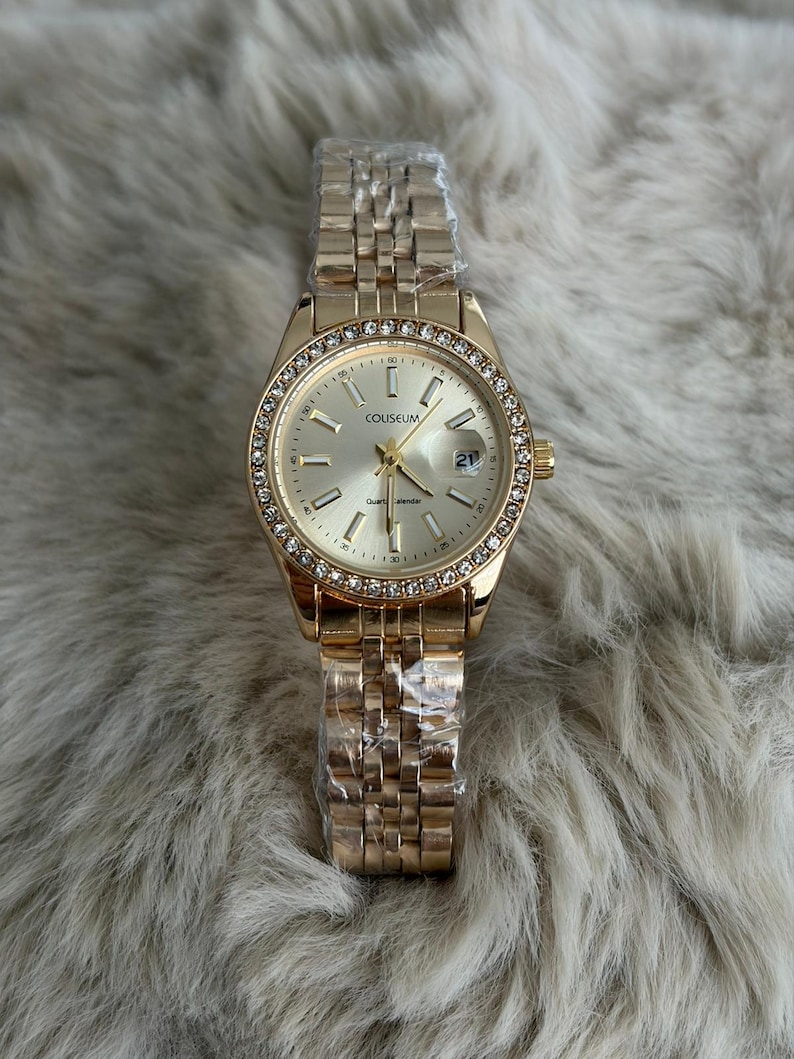 Puede incluir: Reloj de pulsera dorado con esfera redonda y correa met&aacute;lica. La esfera est&aacute; rodeada de peque&ntilde;os cristales transparentes. La esfera del reloj tiene la palabra "COLISEUM" impresa. El reloj a&uacute;n est&aacute; envuelto en pl&aacute;stico protector.
