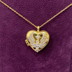 May include: Gold-toned heart-shaped locket pendant necklace. The locket features a detailed bird design with crystal accents. The necklace has a delicate chain and is displayed on a purple background.