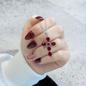 Può includere: Una collana d'argento con un ciondolo a forma di croce con pietre preziose rosse. La croce è composta da cinque pietre preziose rosse a forma di pera.