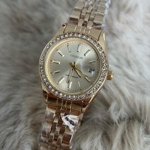 Puede incluir: Reloj de cuarzo Coliseum dorado con esfera redonda y bisel incrustado con peque&ntilde;os cristales transparentes. El reloj tiene una correa dorada y una pantalla de fecha a las 3 en punto. El reloj es nuevo y a&uacute;n tiene el pl&aacute;stico protector en la correa.