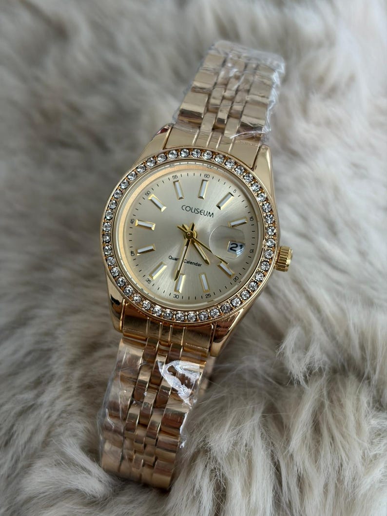 Puede incluir: Reloj de cuarzo Coliseum dorado con esfera redonda y bisel incrustado con peque&ntilde;os cristales transparentes. El reloj tiene una correa dorada y una pantalla de fecha a las 3 en punto. El reloj es nuevo y a&uacute;n tiene el pl&aacute;stico protector en la correa.