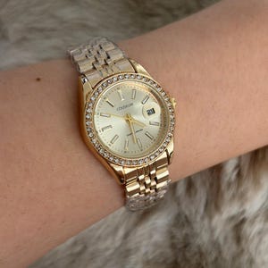 Puede incluir: Reloj de pulsera dorado con esfera clara y visualizaci&oacute;n de la fecha. El reloj presenta un bisel adornado con peque&ntilde;os cristales transparentes y una correa de eslabones dorados. La esfera del reloj muestra la palabra "COLISEUM".