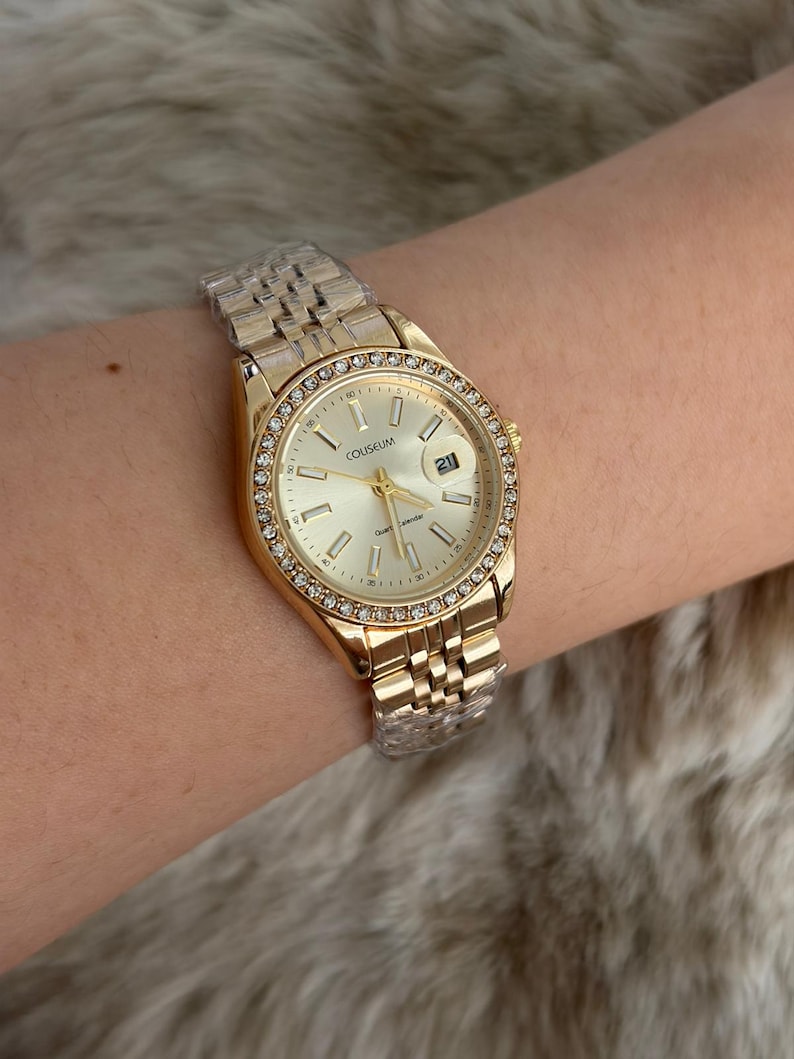 Puede incluir: Reloj de pulsera dorado con esfera clara y visualizaci&oacute;n de la fecha. El reloj presenta un bisel adornado con peque&ntilde;os cristales transparentes y una correa de eslabones dorados. La esfera del reloj muestra la palabra "COLISEUM".