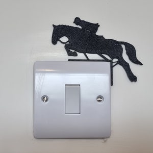 Könnte beinhalten: Ein weißer Lichtschalter mit einer schwarzen Silhouette eines springenden Pferdes mit Reiter. Die Silhouette befindet sich über dem Schalter.