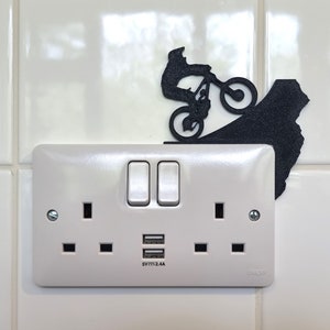 May include: A white electrical socket with two switches and USB ports, with a black silhouette of a mountain biker. The socket is mounted on a white tiled wall.