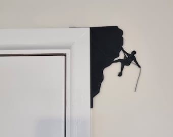 Puede incluir: Silueta negra de un escalador ascendiendo una pared rocosa, montada en un marco de puerta blanco.