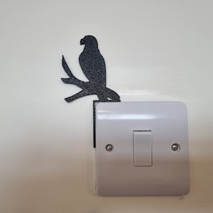 Puede incluir: Una silueta negra de un pájaro posado en una rama, montada en una placa de interruptor de luz blanca.