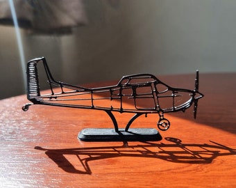 May include: A black wireframe model of a small airplane on a black stand. The model is made of thin black wire and is detailed with a propeller and landing gear.