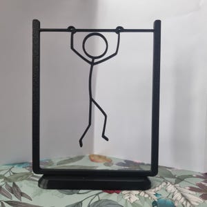 Pode incluir: Escultura decorativa em metal preto com uma figura de palito pendurada numa barra horizontal. A figura está dentro de uma moldura retangular com uma base sólida. Design minimalista, adequado para decoração de casa.