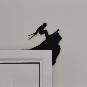 Op de afbeelding: Zwart metalen silhouet van een ski&euml;r in de lucht, geplaatst op de hoek van een wit deurkozijn. De ski&euml;r is afgebeeld met ski's en stokken, tegen een witte muur.