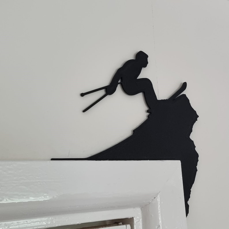 Op de afbeelding: Zwart metalen silhouet van een ski&euml;r in actie, halverwege een sprong. De ski&euml;r wordt afgebeeld met ski's en stokken, tegen een bergachtige vorm. Het kunstwerk is gemonteerd op een wit deurkozijn.