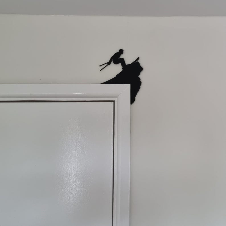 Op de afbeelding: Zwart metalen silhouet van een ski&euml;r in de lucht, geplaatst op de hoek van een wit deurkozijn. De ski&euml;r is afgebeeld met ski's en stokken, tegen een witte muur.