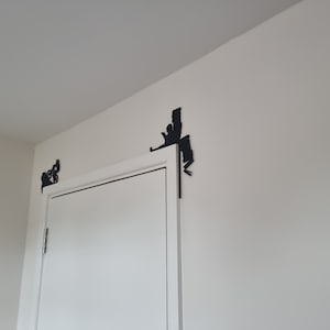 May include: A white door with two black metal door stops in the shape of a person climbing a wall. The door stops are attached to the wall above the door.