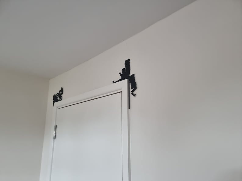 May include: A white door with two black metal door stops in the shape of a person climbing a wall. The door stops are attached to the wall above the door.