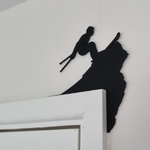 Op de afbeelding: Een zwart metalen wandkunstwerk dat een ski&euml;r in actie toont. Het silhouet toont een ski&euml;r in de lucht, met ski's en stokken, tegen een witte muur. Het kunstwerk is bevestigd op de hoek van een wit deurkozijn.