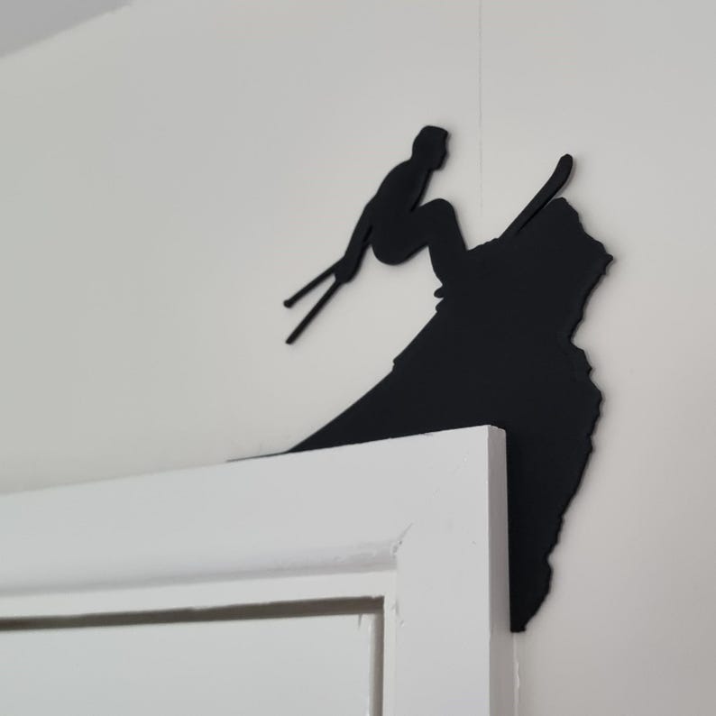 Op de afbeelding: Een zwart metalen wandkunstwerk dat een ski&euml;r in actie toont. Het silhouet toont een ski&euml;r in de lucht, met ski's en stokken, tegen een witte muur. Het kunstwerk is bevestigd op de hoek van een wit deurkozijn.