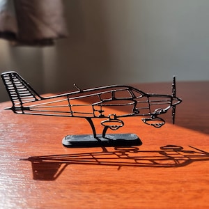 Puede incluir: Un modelo de avión pequeño de alambre negro en un soporte negro. El modelo está sobre una superficie de madera.