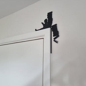 May include: Black silhouette of a person reading a book, with a stack of books behind them, mounted on a white wall above a white door.