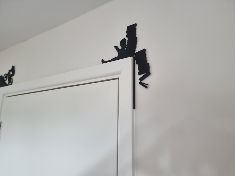 May include: Black silhouette of a person reading a book, with a stack of books behind them, mounted on a white wall above a white door.