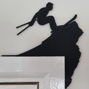 Op de afbeelding: Zwarte metalen wandkunst met een ski&euml;r in actie, met ski's en stokken. De ski&euml;r bevindt zich boven een bergvormig silhouet. De wandkunst is gemonteerd op een wit oppervlak.