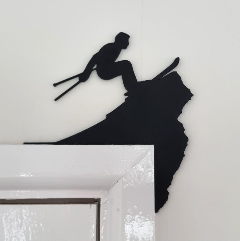 Op de afbeelding: Zwart metalen wandkunstwerk dat een ski&euml;r in actie toont, in de lucht boven een berg. Het silhouet is gemonteerd op een witte muur, met de hoek van een wit deurkozijn op de voorgrond.