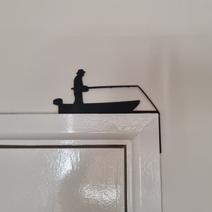 Peut inclure: Silhouette noire en métal d'une personne pêchant depuis un bateau, montée sur un cadre de porte blanc.