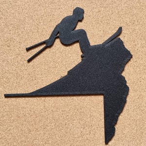 Op de afbeelding: Zwart metalen silhouet van een ski&euml;r in actie, tegen een getextureerde achtergrond. De ski&euml;r is in de lucht, met ski's en stokken zichtbaar. Het ontwerp bevat een gestileerde bergvorm, wat een dynamische en sportieve uitstraling cre&euml;ert.