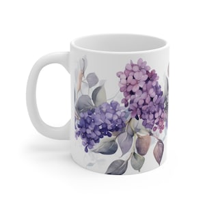 May include: White ceramic mug with a floral design featuring purple and pink flowers and green leaves. The flowers are watercolor style and are arranged in a horizontal band around the mug.